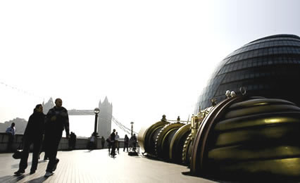 A panaoramic view of the Telectroscope, The Mayor’s office and London Bridge in silhouette. A couple are glancing at the device as the walk past.